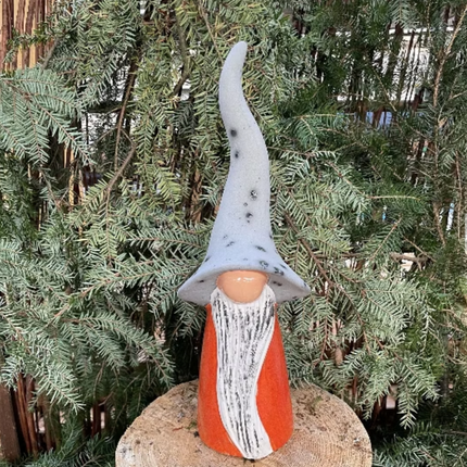Gnome figurine with a long gray hat and orange and white striped outfit standing on a rock with green foliage in the background.