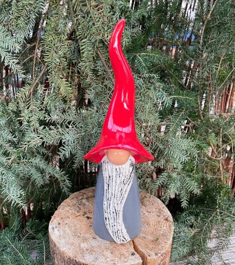 Lifestyle picture showing a grey gnome sitting on a log with foliage of an evergreen tree as a backdrop from Goose and Company.