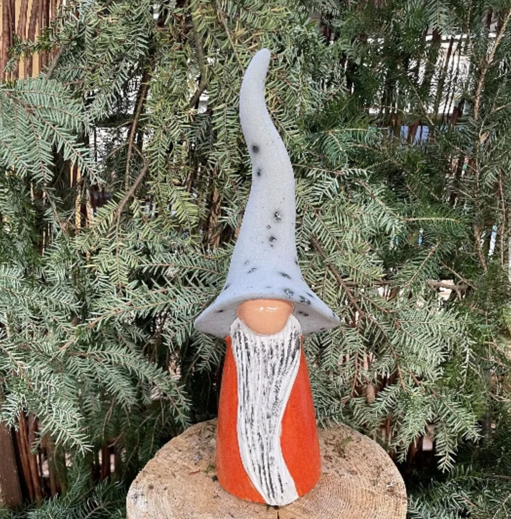 Gnome figurine with a long gray hat and orange and white striped outfit standing on a rock with green foliage in the background.